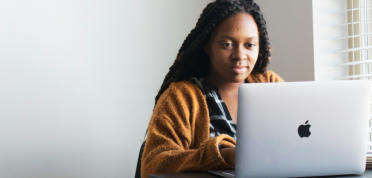 A young woman in a sweater is sitting at a desk looking at a laptop.